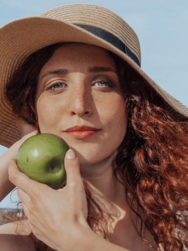 A relaxed woman with a hat holds a green apple, enjoying a sunny beach day.