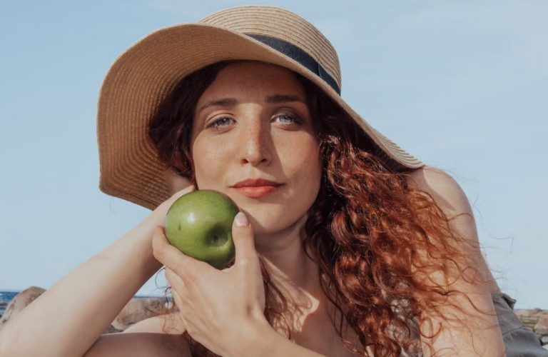 A relaxed woman with a hat holds a green apple, enjoying a sunny beach day.