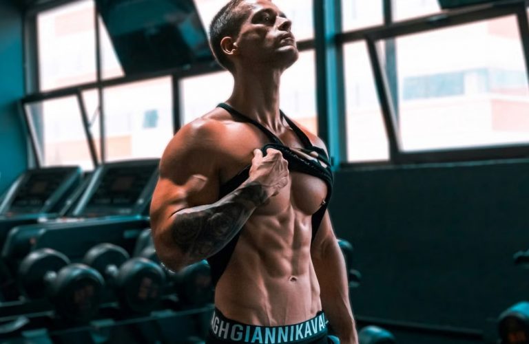 Muscular man showing abs and tattoos in a gym setting.